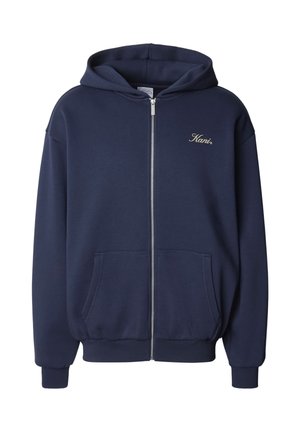 Tumši zila, ar rāvējslēdzēju aizverama kapuce, izgatavota no mīkstas auduma. Iespējas ietver priekšējo kabatu, ribotās aproces un mazu zelta logotipu kreisajā krūtī.