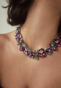 Mehrfarbige Edelsteinkette mit pinken, blauen und lila Steinen in verschiedenen Formen, ergänzt durch goldfarbene Metallelemente. Funkelnde Details sind sichtbar.