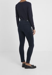 Jeans skinny azul oscuro de mezclilla, con cintura alta, bolsillos traseros y dobladillo deshilachado. Combinados con zapatos planos negros.