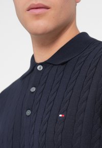 Granatowa dziana koszulka polo z wzorem w warkocze, z trzema guzikami i kołnierzykiem. Posiada małe logo w kolorach czerwonym, białym i niebieskim po lewej stronie.