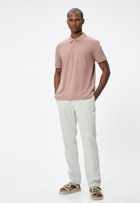 Polohemd aus leichtem rosa Baumwollstoff mit texturiertem Kragen, kombiniert mit hellbeigen Leinenhosen und tanfarbenen Sandalen mit geflochtenen Akzenten.