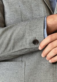 Sivkasta blazer tkanina v ribjem vzorcu; z značilnim črnim gumbom na manšeti in modro srajco pod njim.