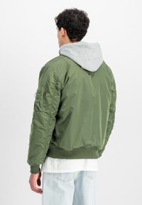 Giacca bomber verde con fodera in hoodie grigia, polsini e orlo a coste, e tessuto texture. Presenta una tasca sulla manica sinistra.