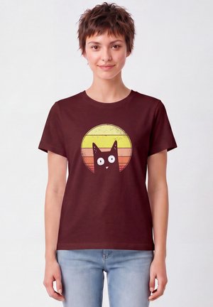 Femme aux cheveux courts et bruns portant un t-shirt bordeaux avec un chat noir de dessin animé et un fond circulaire jaune-orange.
