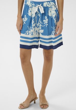 Pantaloni scurți cu imprimeu floral alb și albastru cu dungi orizontale, talie cu șnur, purtați cu sandale bej cu toc și degetele goale pe o persoană în picioare.