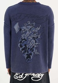 Navy-blaues Rippenpullover mit langen Ärmeln, der einen grafischen Druck auf der Rückseite aufweist. Der Stoff hat ein strukturiertes Aussehen mit kontrastierenden Akzenten.