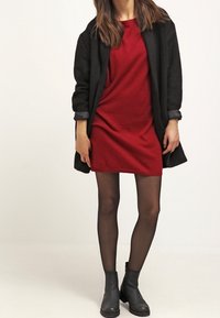 Robe tricotée rouge avec un manteau noir ample, des bottines noires et des collants noirs transparents. Le manteau a une finition texturée et des manches retroussées.