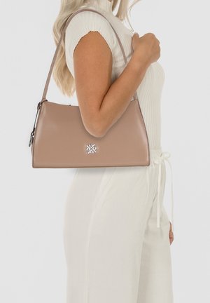 Beige-farbenes Kunstleder-Handtäschchen in dreieckiger Form, mit silbernem Logo-Detail und einem gebogenen Tragegurt, gehalten von einer Person, die ein hellfarbiges Outfit trägt.