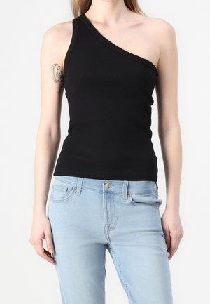 Mujer con un top negro de un solo hombro y vaqueros azul claro, mostrando un tatuaje en su brazo izquierdo superior.