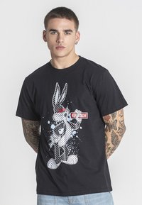 Černé bavlněné tričko s grafikou karikatury králíka s červenými akcenty, umístěné na pozadí s rozstřikovaným designem v různých odstínech modré.