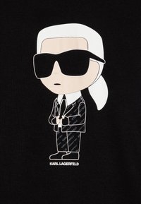 Zwart stof met een cartoonfiguur met wit haar, een zonnebril en een zwart pak. "Karl Lagerfeld" staat eronder weergegeven.