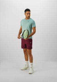 Mann in türkisem Shirt, maronen Shorts, weißen Schuhen, der einen Tennisschläger hält und vor einem schlichten hellgrauen Hintergrund steht. Er blickt nach links.