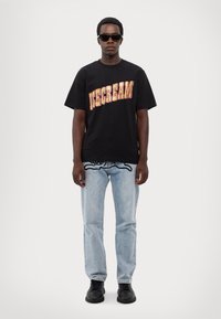 Black t-shirt featuring bold, colorful "ICECREAM" text, paired with light blue jeans and black shoes. Model wearing sunglasses.
