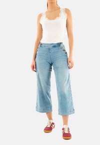Pantalones de denim de pierna ancha en azul claro con botones laterales y un dobladillo deshilachado, combinados con una camiseta sin mangas con encaje blanco y zapatillas burdeos.