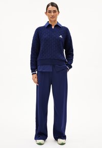 Marineblauer Strickpullover mit V-Ausschnitt und gerippten Bündchen, über einem marineblauen Hemd getragen, kombiniert mit locker sitzenden marineblauen Hosen und weißen Sneakers.