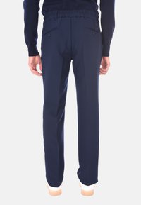 Uomo che indossa pantaloni su misura blu navy con tasche posteriori con bottone, abbinati a sneaker bianche e a un top lungo scuro, in piedi di spalle.