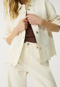 Ivoorkleurig spijkerjack met een knoopsluiting aan de voorkant en oogjesdetails aan de zoom, gecombineerd met lichtgekleurde broek en een bruin topje eronder.
