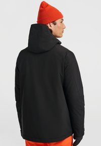 Zwarte gevoerde jas met capuchon, voorzien van een gladde textuur. Draagt een oranje beanie, wat een sportief ontwerp en praktische stijl laat zien.