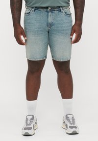 Shorts in denim bluastro con taglio dritto, design a cinque tasche e texture sbiadita. Indossati con sneaker grigie e calzini bianchi alla caviglia.