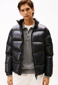 Black puffer jacket with a shiny finish, quilted design, high collar, and zippered front. Worn over a gray cable-knit sweater.