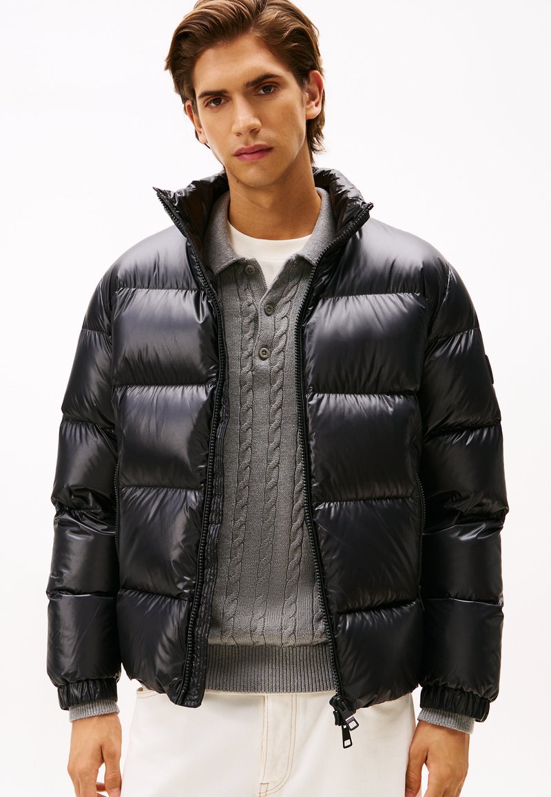 Black puffer jacket with a shiny finish, quilted design, high collar, and zippered front. Worn over a gray cable-knit sweater.