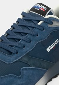 Μπλε αθλητικό παπούτσι με υφασμένο πλέγμα και επάνω μέρος από σουέντ, μαλακό κολάρο και επίπεδα κορδόνια· διαθέτει λευκό λογότυπο και μαύρες/γκρι λεπτομέρειες.