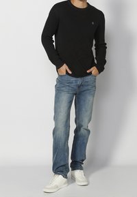 Muž v černé dlouhé košili, modrých džínech a bílých teniskách, stojící s rukama v kapsách na čistém bílém pozadí.