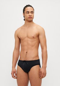 Schwarze Schwimmshorts mit einem Kordelzug, die ein glattes, dehnbares Material und ein enges Design aufweisen, getragen von einem männlichen Model vor einem schlichten Hintergrund.