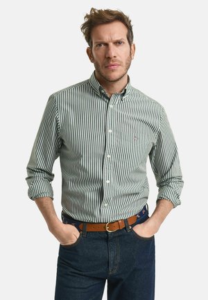 Mann mit hochgekrempelten Ärmeln, trägt ein grün-weiß gestreiftes Hemd, dunkle Jeans und einen braunen Gürtel, die Hände in den Taschen.