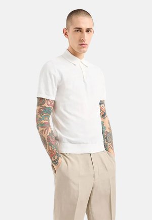 Polo bianca realizzata in tessuto a maglia con colletto a coste e maniche corte, indossata con pantaloni beige. Tatuaggi visibili sulle braccia.