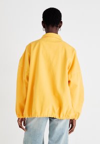 Gul oversized jakke med rund krage, elastisk kant og vide ermer, kombinert med lyseblå denimjeans.