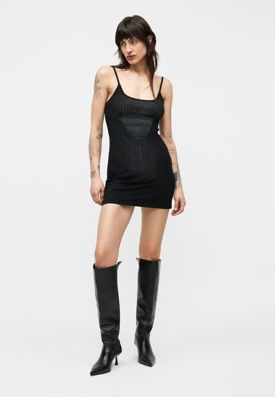 Γυναίκα με τατουάζ που φοράει εφαρμοστό μαύρο μίνι φόρεμα και μαύρες μπότες με τακούνι μέχρι το γόνατο, στέκεται μπροστά σε απλό φωτεινό φόντο.