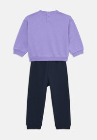 Paarse sweatshirt met drie knopen op de rug en marineblauwe broek. Beide items hebben een zachte textuur en een losse pasvorm.