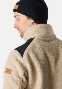 Beige fleece jacket with black shoulder accents, high collar, and leather label on the left sleeve. Fabric has a textured, soft finish.