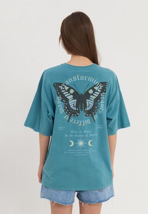 Mujer con cabello largo castaño lleva una camiseta azul verdosa de gran tamaño con una gran mariposa negra y azul y un texto sobre la transformación en la parte trasera.