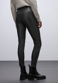 Leggings de cuero sintético negro con una textura suave y un ajuste ceñido, combinados con botines negros, exhibidos sobre un fondo neutro.