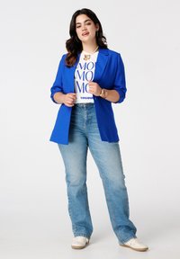 Blazer bleu sur un t-shirt graphique blanc avec du texte bleu, associé à un jean flare délavé et des baskets blanches. Bijoux dorés en accent.