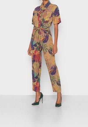 Femme portant une combinaison à boutons, manches courtes, avec des motifs colorés de feuilles tropicales et une ceinture nouée à la taille, assortie à des talons hauts verts.