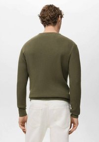 Olivgrüner, gerippter Pullover mit rundem Ausschnitt und langen Ärmeln, der eine taillierte Passform und einen strukturierten Stoff aufweist. Kombiniert mit weißen Hosen.