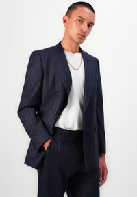 Marineblå stribede jakkesæt med en dobbeltradet blazer, kombineret med en hvid t-shirt og en guldkæde, med slanke klapper og skræddersyet pasform.
