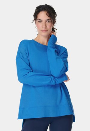 Vrouw met donker krullend haar draagt een blauwe trui met lange mouwen en donkere broek, poseert met één hand bij haar nek en gekruiste armen.