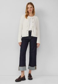 Gilet en tricot blanc avec poches, associé à un jean large et foncé avec des ourlets retroussés. Le modèle porte des mocassins bruns. Design simple.