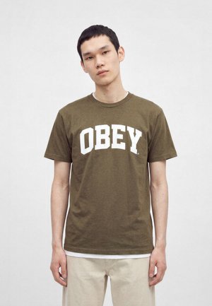 Νέος άνδρας που φοράει μπλουζάκι ελιάς με "OBEY" σε μεγάλα λευκά γράμματα και μπεζ παντελόνι, μπροστά σε απλό φωτεινό φόντο.