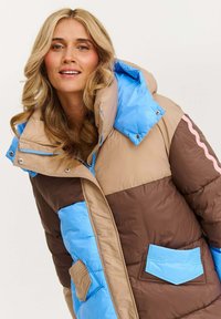Žena s blond vlasmi, ktorá má na sebe pufrovú bundu v kombinácii hnedé, béžové a modré farby, s veľkým golierom a prednými vreckami, s mierne úsmevom.
