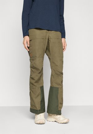 Person wearing olive green cargo pants with reinforced dark green patches on lower legs and beige hiking shoes, standing against a plain background.
