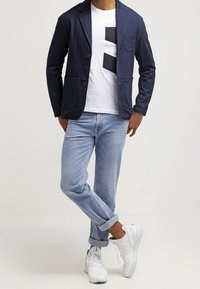 Blazer de algodón azul marino con dos bolsillos delanteros, camiseta blanca con un diseño geométrico negro, vaqueros azul claro y zapatillas blancas.