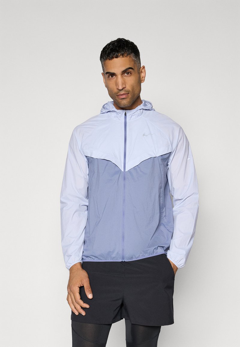 Giacca leggera in blu chiaro e grigio, con zip frontale, cappuccio, polsini elastici e un design geometrico sul petto.