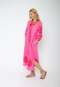 Kobieta stojąca w różowej sukience midi z czerwoną haftowaną florystyką i bocznym rozcięciem, nosząca beżowe płaskie sandały związywane na sznurki na białym tle.