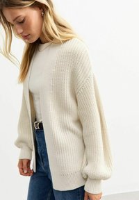 Cardigan lavorato a maglia in bianco sporco, con una vestibilità ampia, texture a costine e maniche larghe. Abbinato a una maglietta attillata color crema e jeans blu.