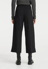 Pantalons larges noirs avec une taille haute, dotés de plis avant et de fentes latérales. Associés à des bottes de combat noires.
